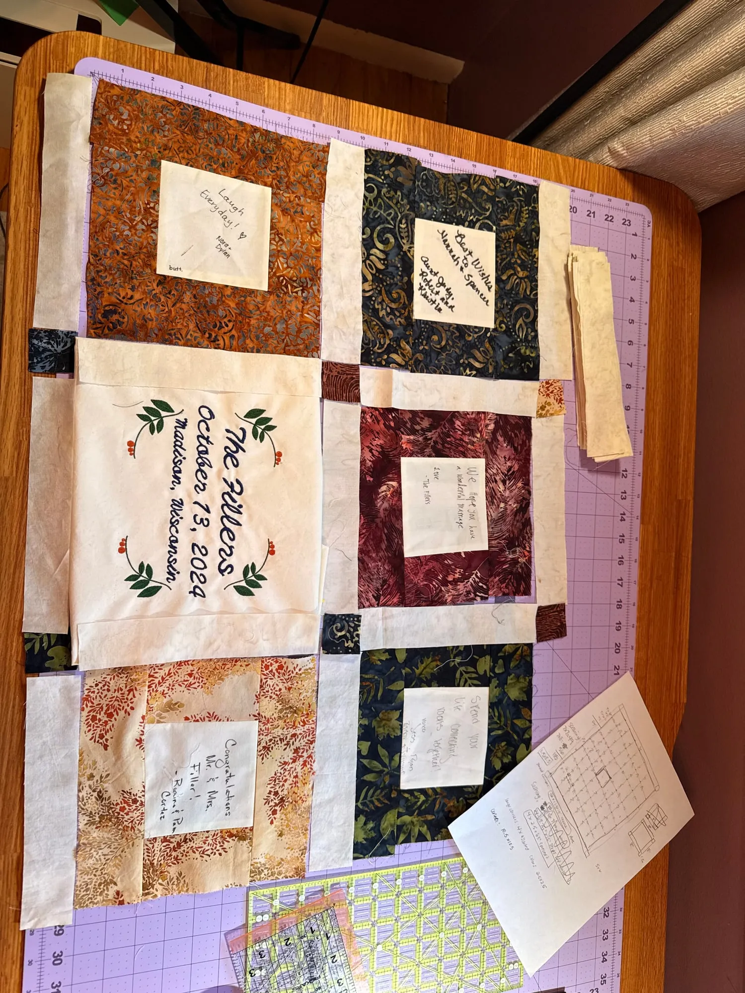 Six pieced blocks laid out on a purple cutting mat, each a cream signed square framed by batik fabric, with a hand-drawn pattern sketch on graph paper beside them.