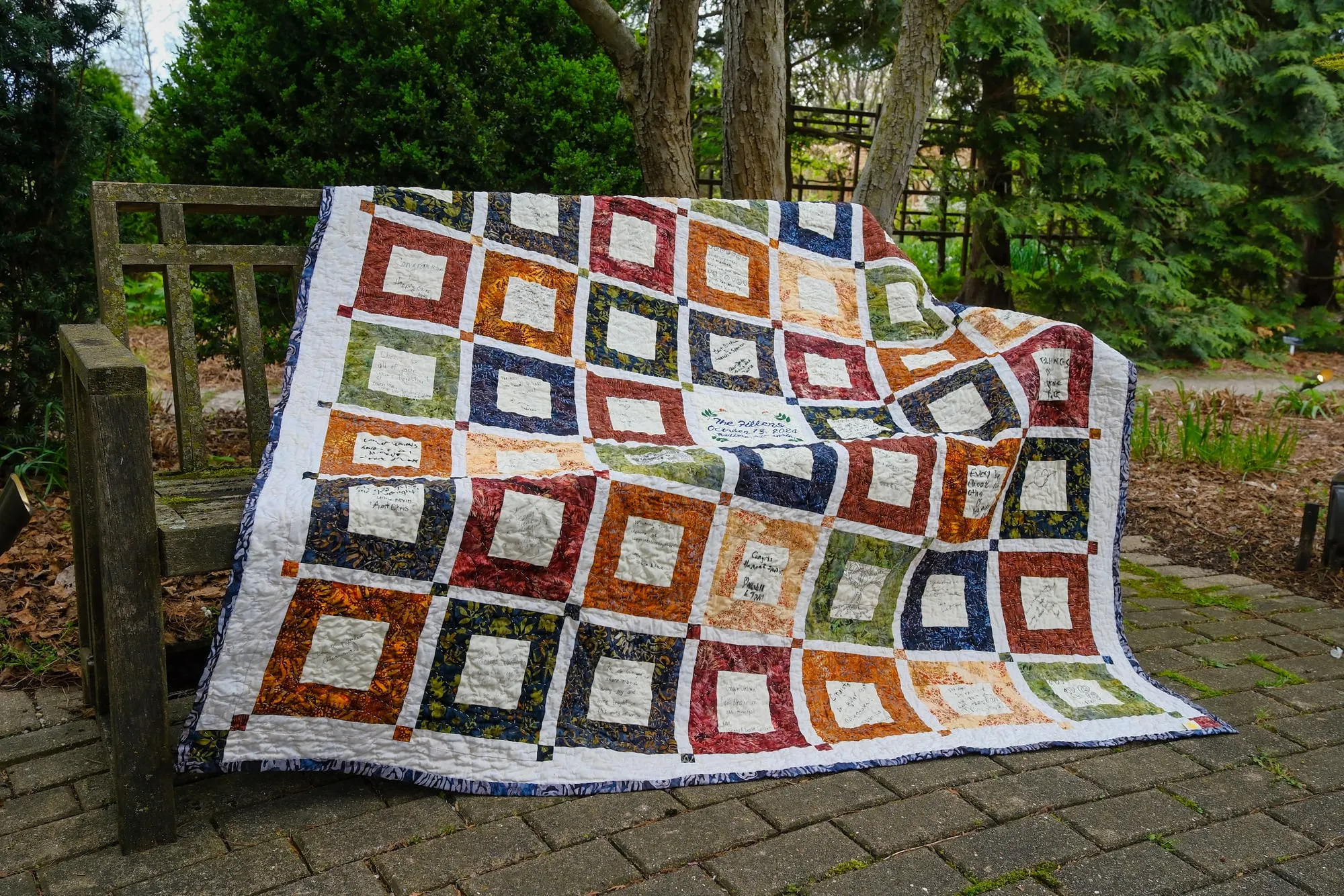 A finished signature quilt draped over a wooden garden bench, made of batik squares in rust, orange, olive and navy, each framing a cream center signed by a wedding guest.