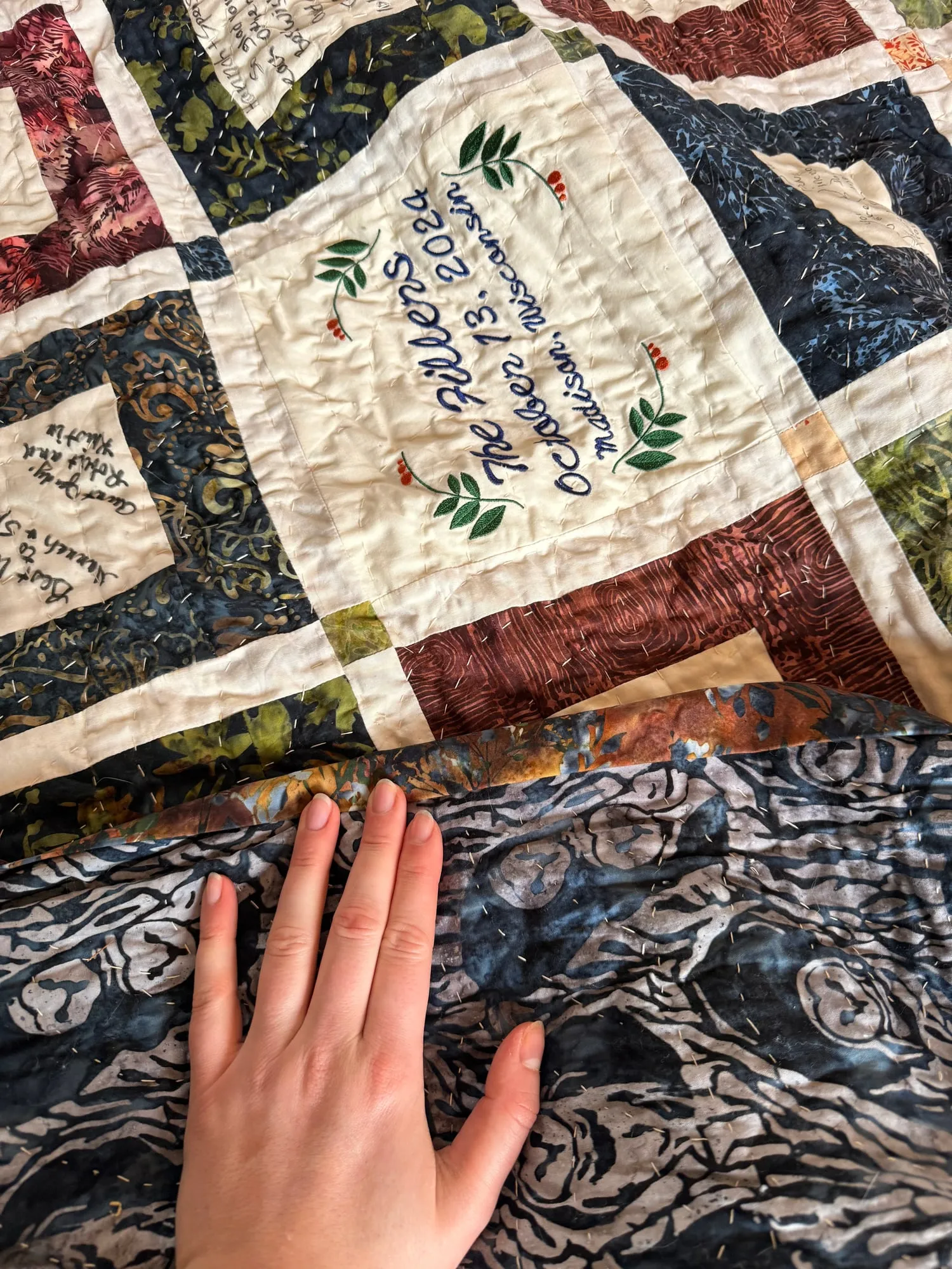 An embroidered label block reading The Fillers, October 13, 2024, Madison, Wisconsin, surrounded by signed squares, with a hand resting on the navy batik backing.